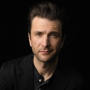 A headshot of M. Scott McLean*. He is a white man, wearing a dark gray button up and a dark black suit jacket. He has short, coifed brown hair, light facial hair, and a half-smile.