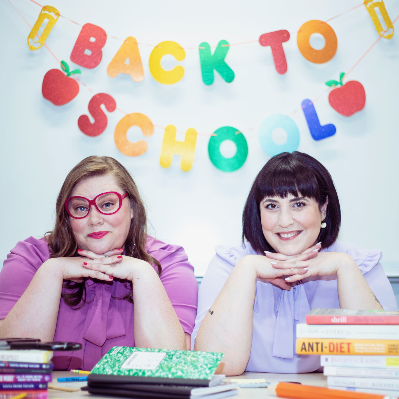 Kelsie Jepsen and Olivia Custodio sit at a desk in front of colorful hanging letters spelling WELCOME TO SCHOOL. Kelsie has long brown hair and wears a mauve blouse with ruffles at the neck and large round red glasses, her nails are also painted red. Olivia wears a similar blouse, but it is a lavender purple, she has dark brown hair cut into a bob that sits at her shoulders. The desk is covered in classroom items: books, journals, pens, pencils and markers.